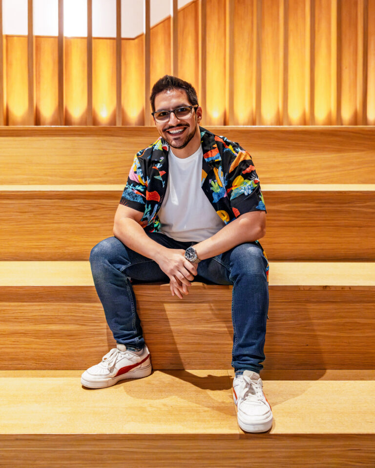 Raphael Pavel sitting on modern wooden tiered seating, smiling at the camera while wearing a colourful patterned shirt, white T-shirt, jeans, and sneakers, with warm backlit wood panels behind him.