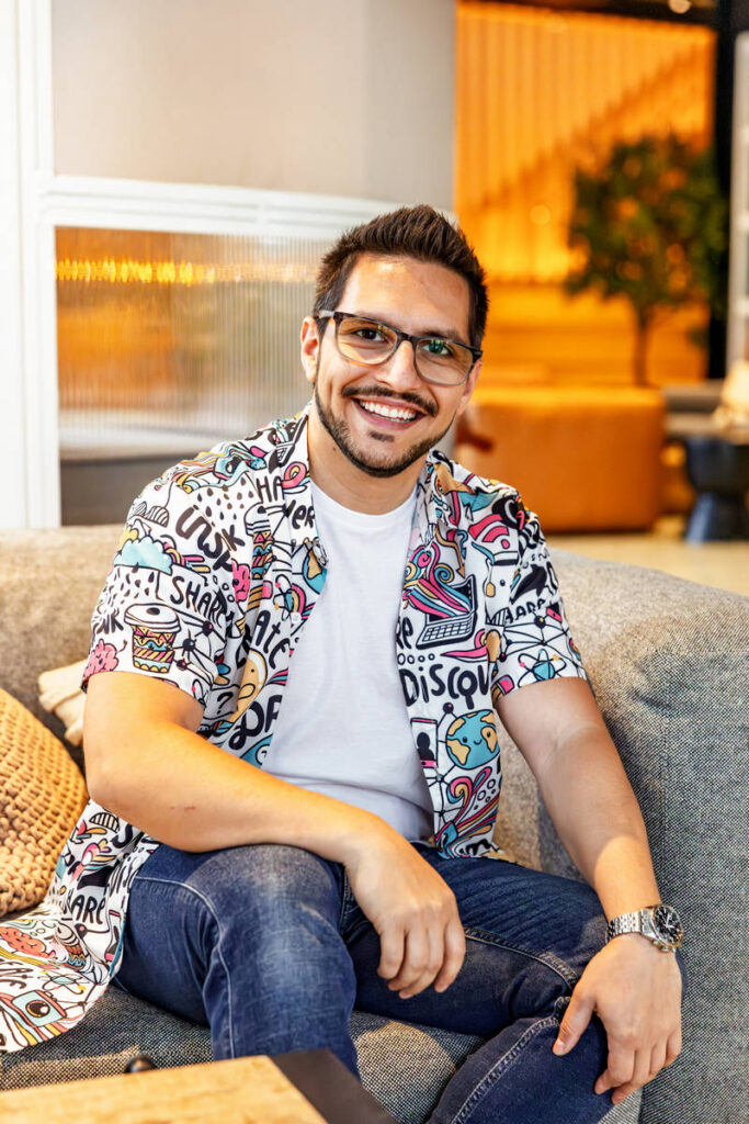 About 2 Raphael Pavel sitting on a modern grey sofa, smiling at the camera while wearing glasses, a white T-shirt, and a colorful patterned shirt with playful graphics, with warm interior lighting and a blurred plant in the background.